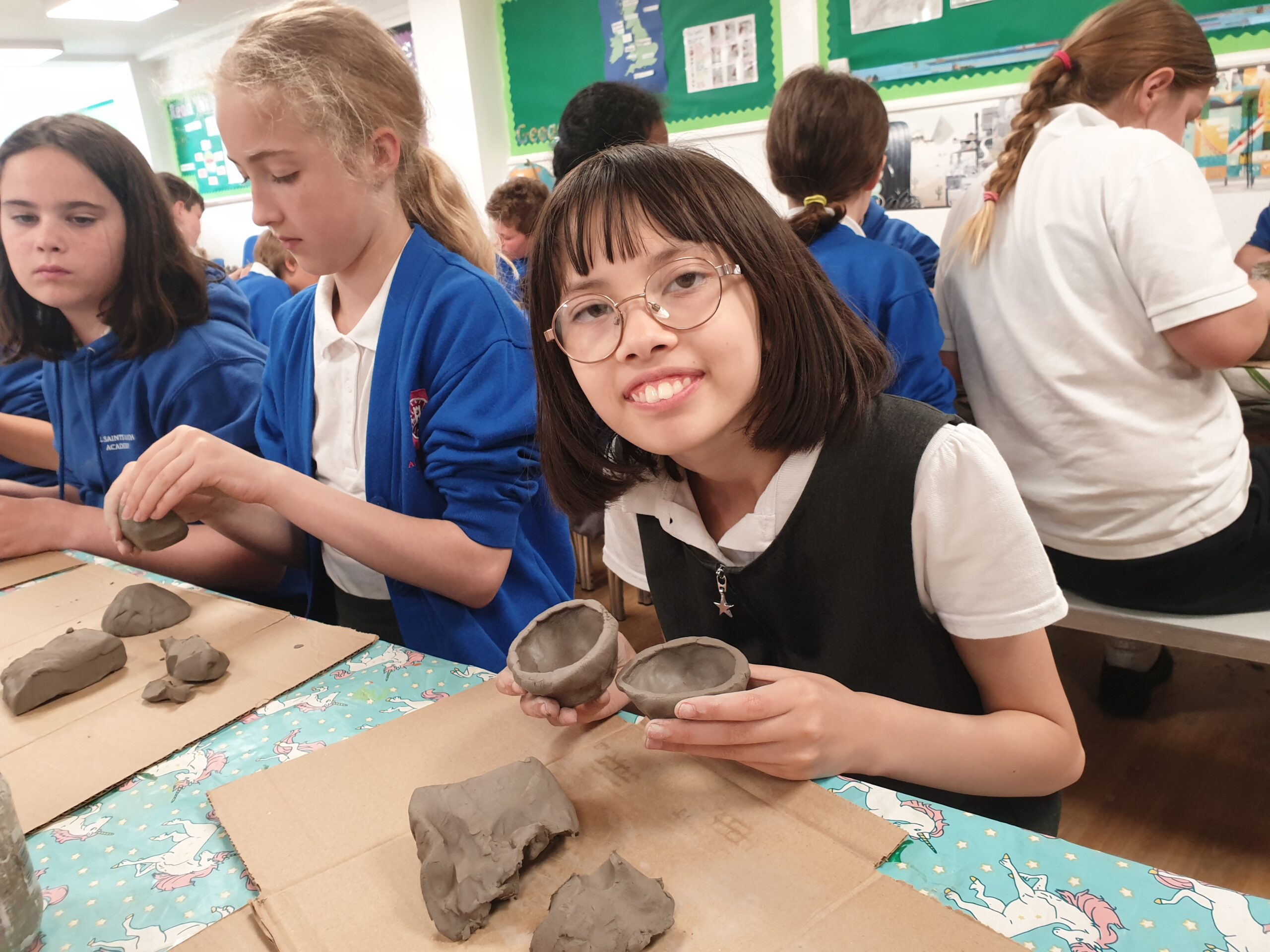 Young girl with glasses in classroom proudly holds up clay pot